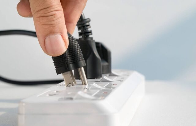 A hand plugging a black, two-prong plug into a white power strip with multiple other plugs connected to the power strip.
