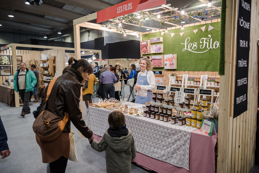 A food stall at a market, with a vendor offering jams and truffles to customers. 